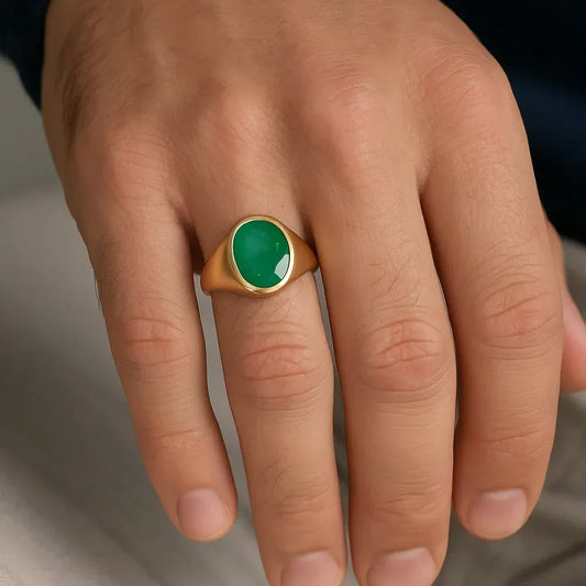 Hand wearing a gold ring with a green emerald stone on a neutral background