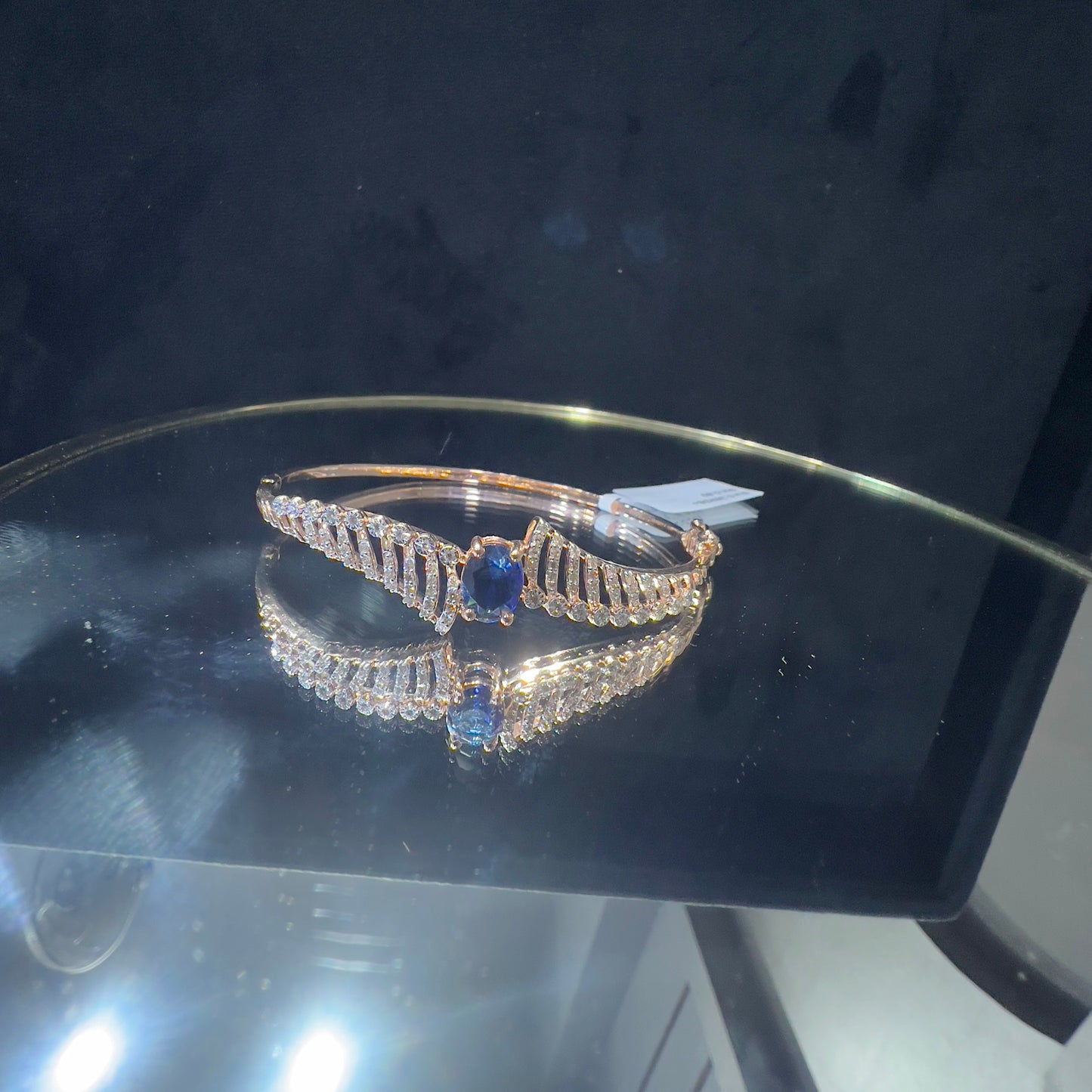 A luxurious gold diamond bracelet with a brilliant cut blue stone in an openable design, displayed on a reflective surface.
