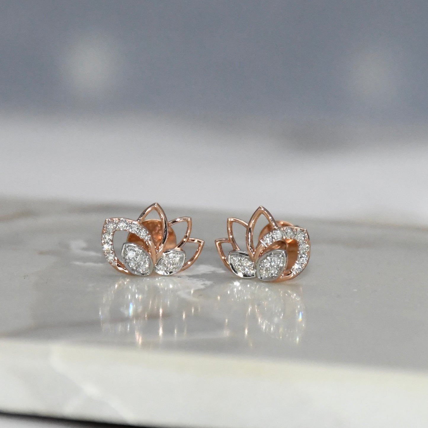 Rose gold lotus flower earrings with clear diamonds on a reflective surface.