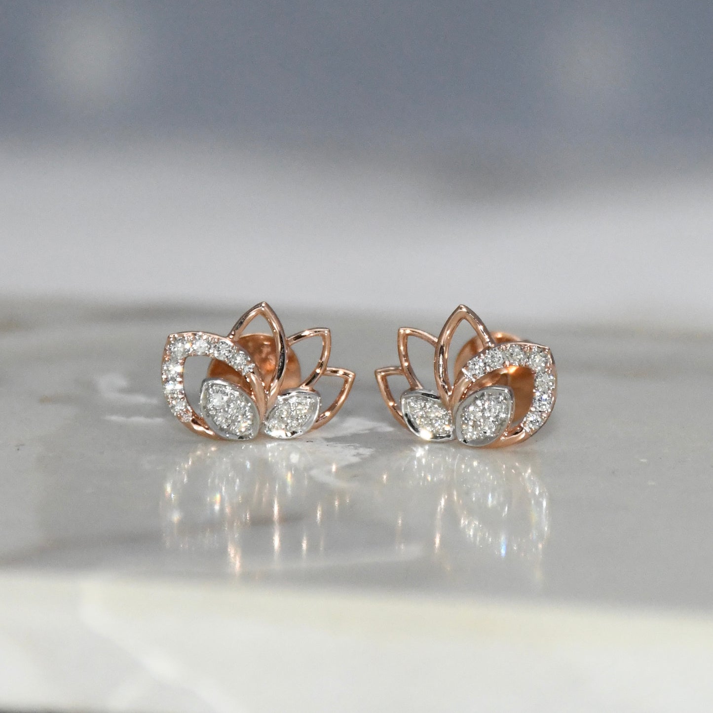 Rose gold lotus flower earrings with clear diamonds on a reflective surface.