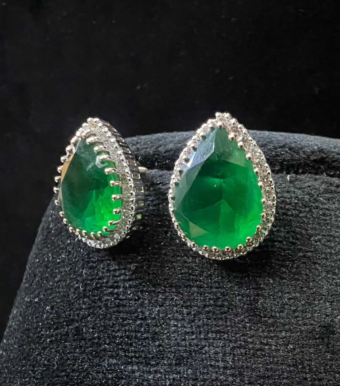 Pair of teardrop green stone and diamond halo earrings displayed on a black stand.
