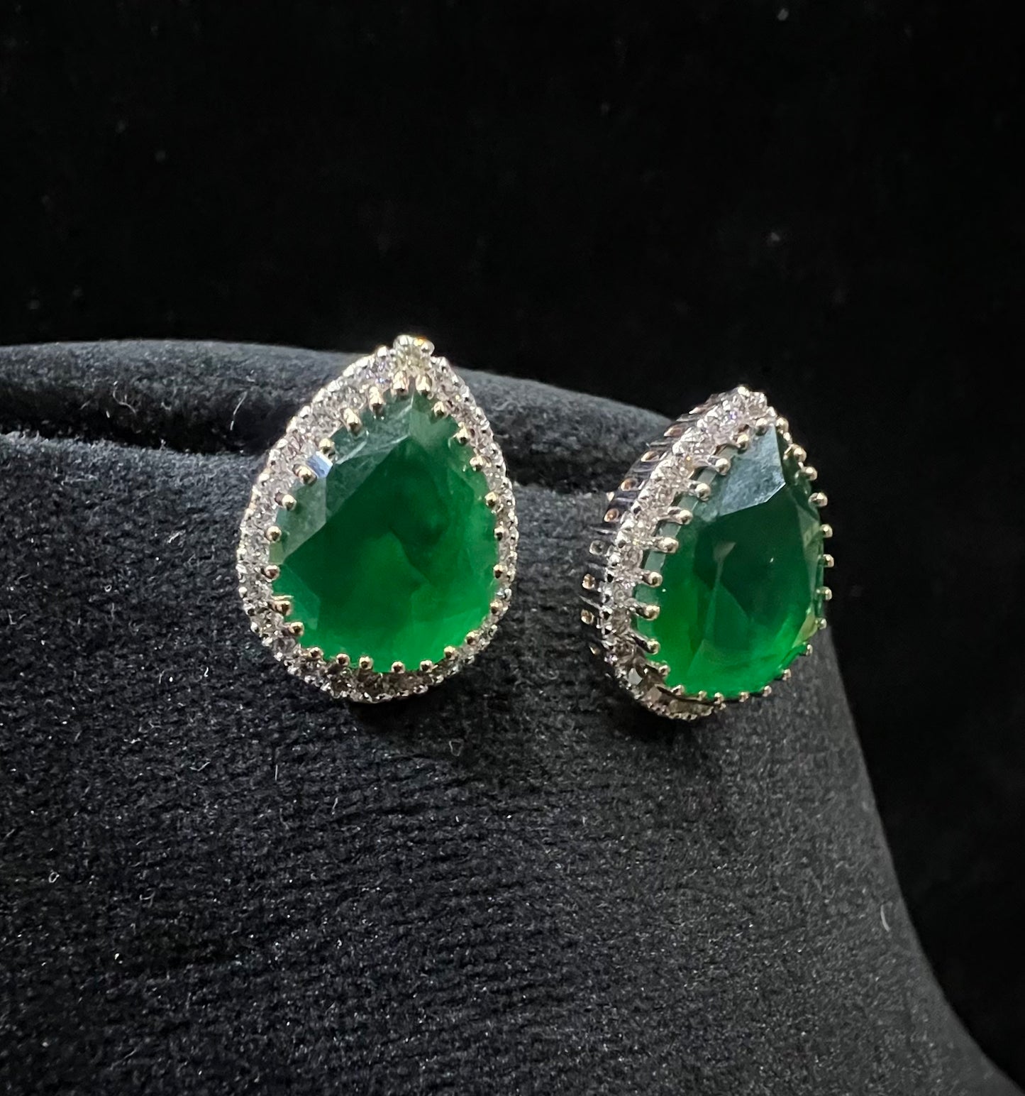 Pair of teardrop green stone and diamond halo earrings displayed on a black stand.