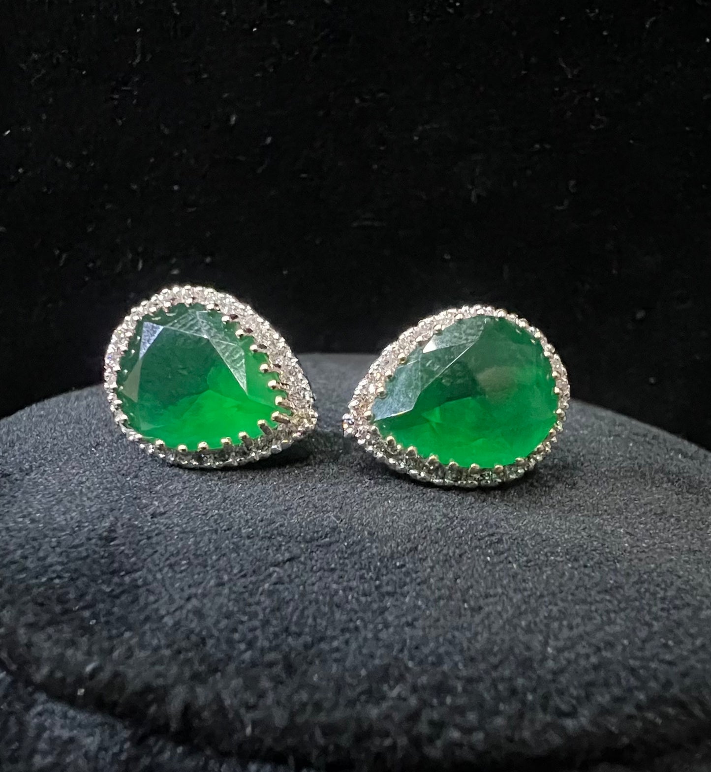 Pair of teardrop green stone and diamond halo earrings displayed on a black stand.