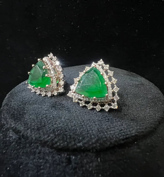 A pair of gold diamond earrings with a green color stone set in a minimalist design on a black background.