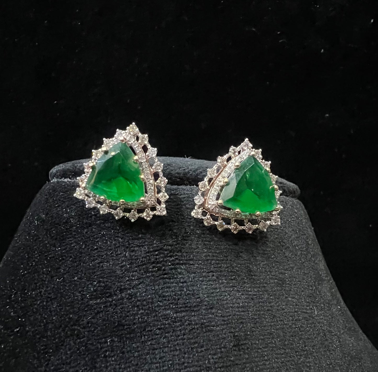 A pair of gold diamond earrings with a green color stone set in a minimalist design on a black background.