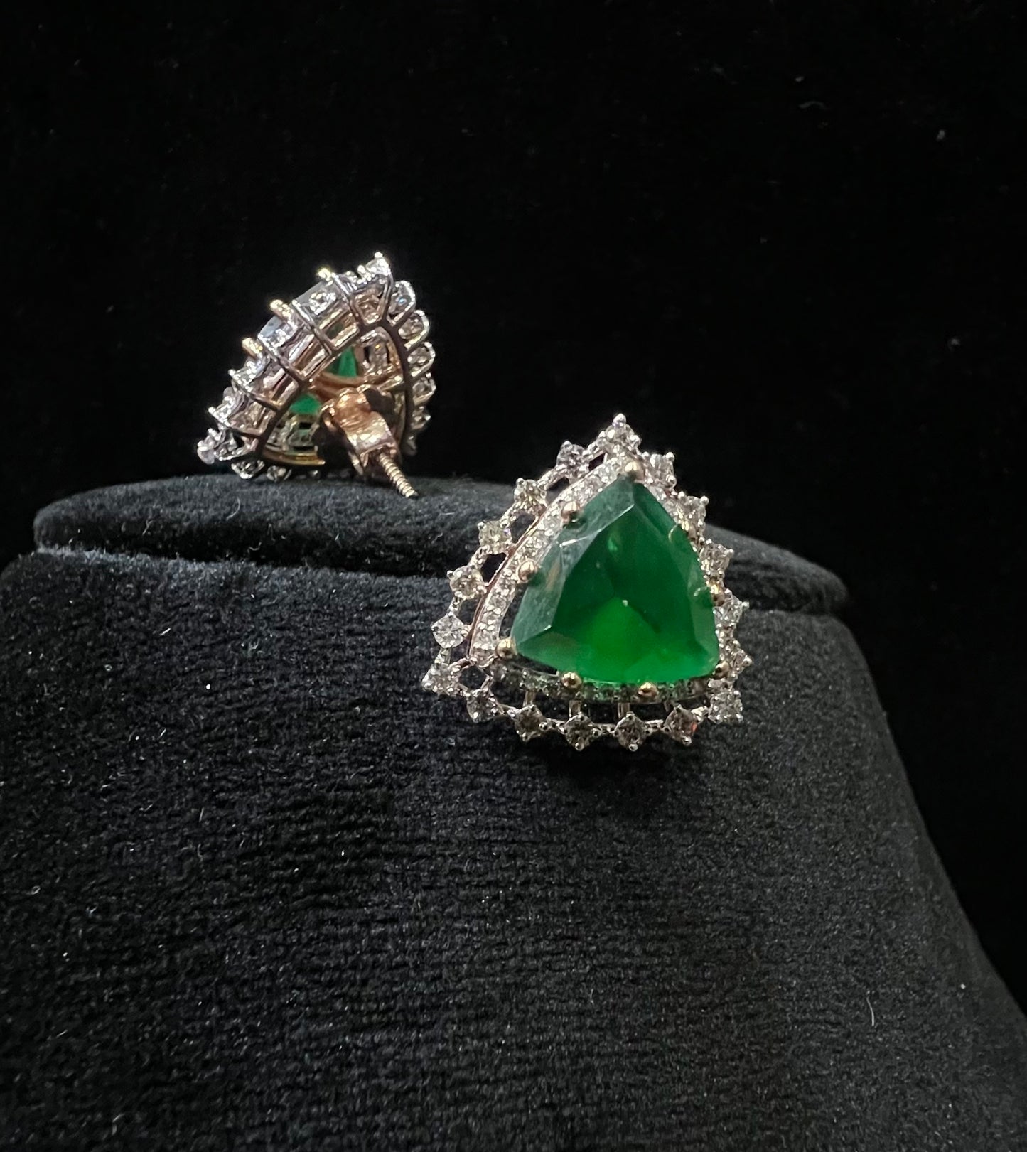 A pair of gold diamond earrings with a green color stone set in a minimalist design on a black background.