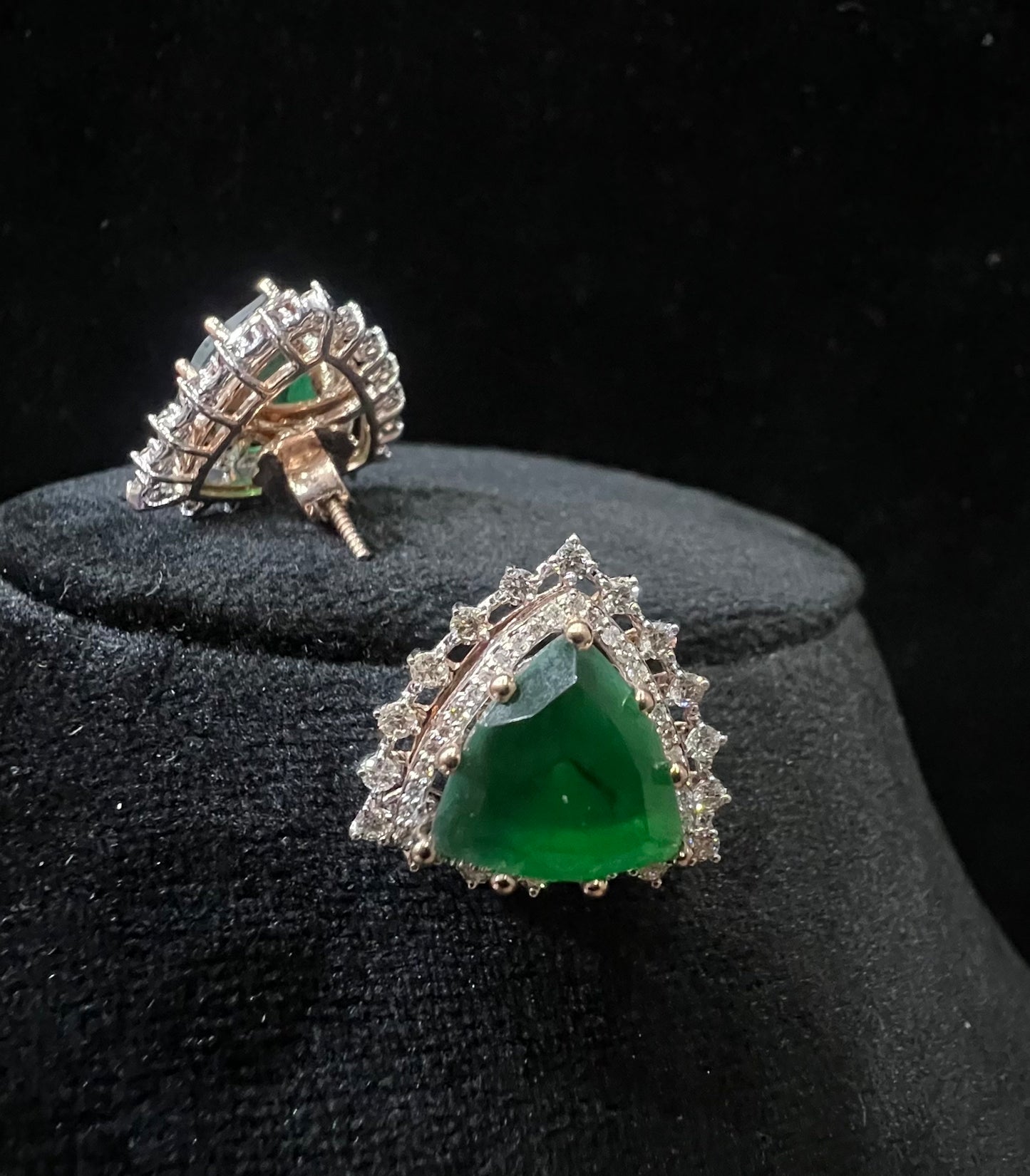 A pair of gold diamond earrings with a green color stone set in a minimalist design on a black background.