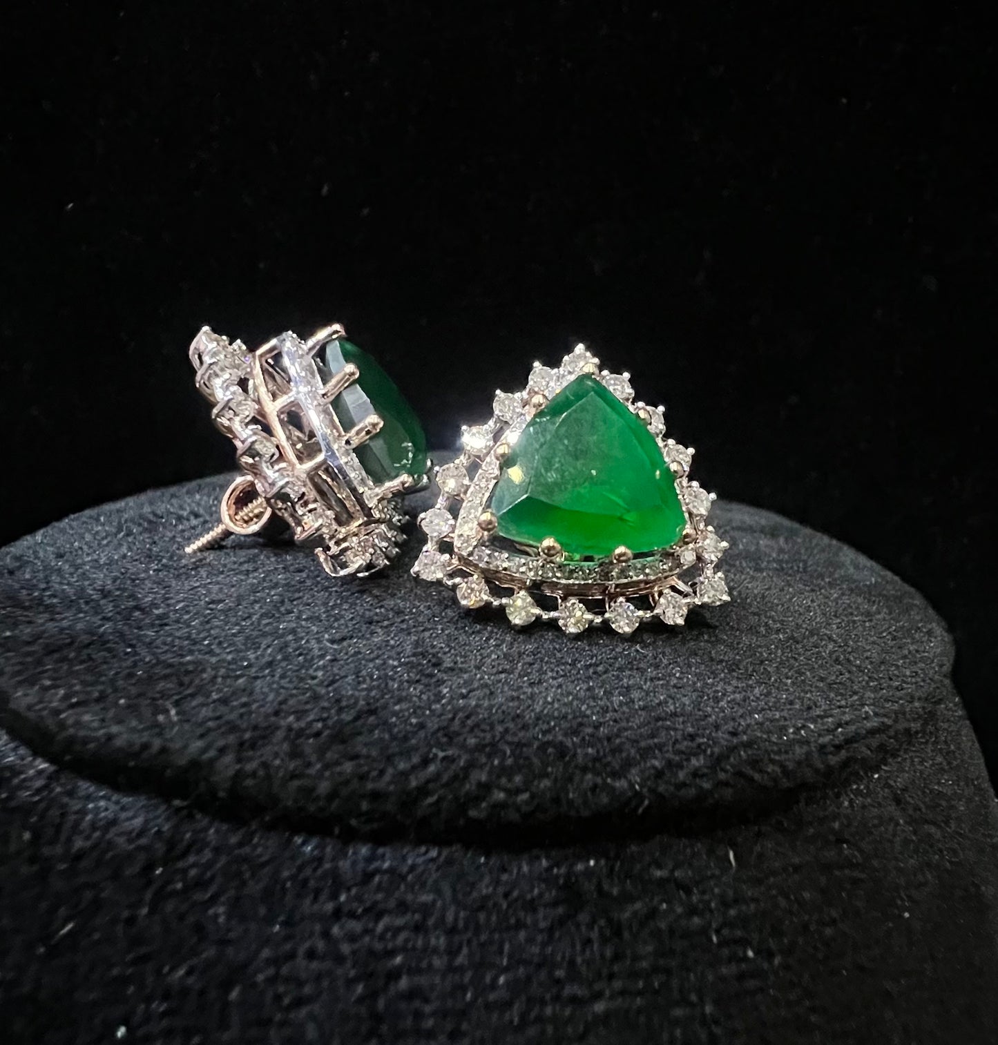 A pair of gold diamond earrings with a green color stone set in a minimalist design on a black background.