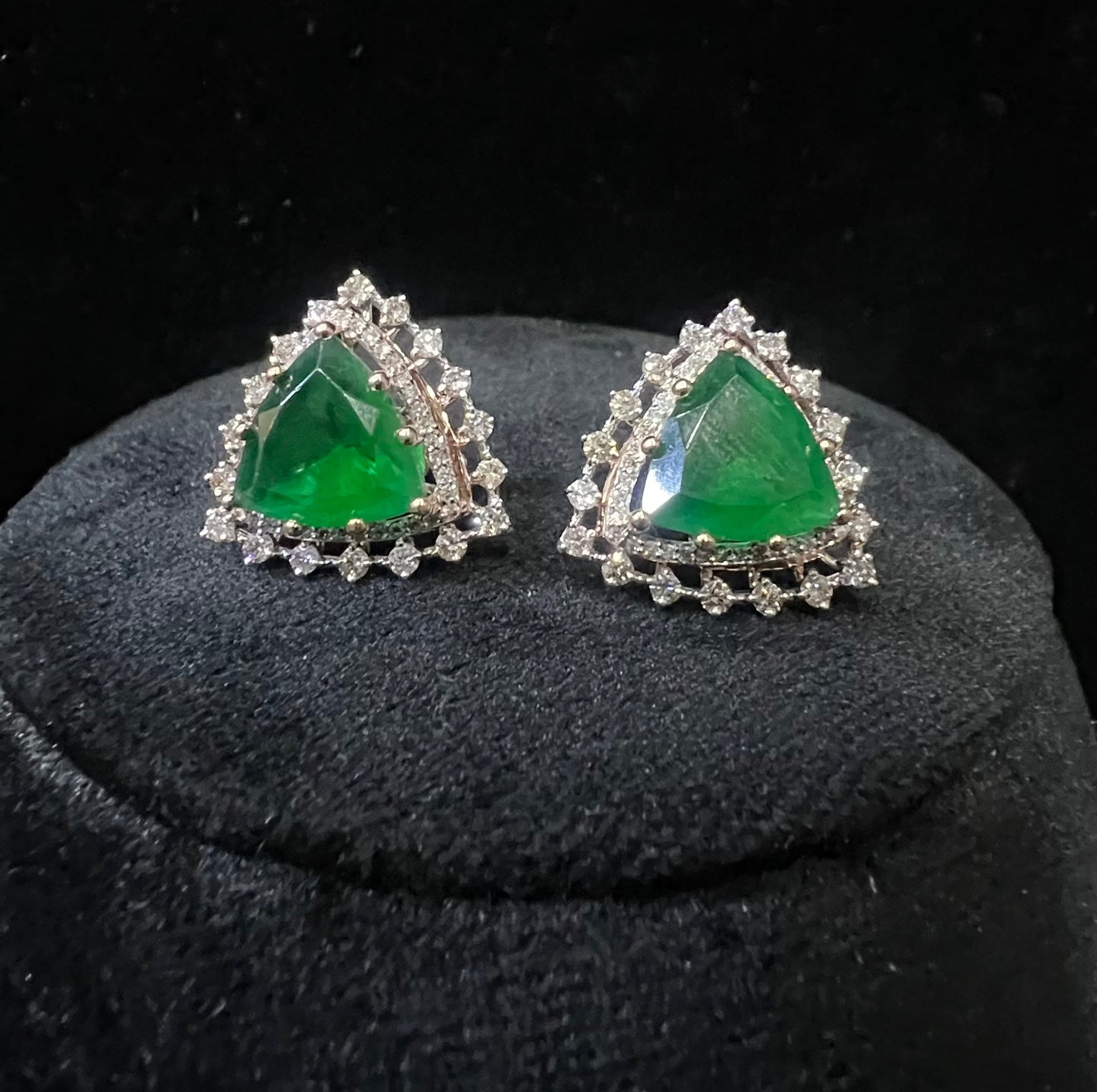 A pair of gold diamond earrings with a green color stone set in a minimalist design on a black background.