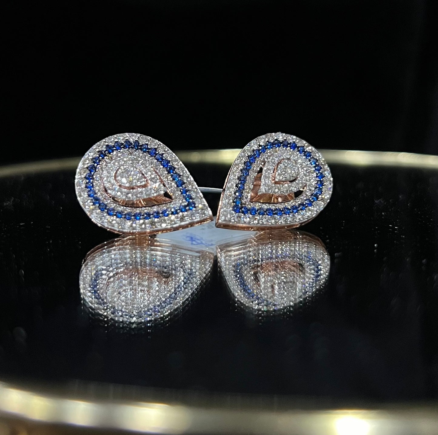 A pair of gold diamond earrings with a geometric pattern, displayed on a reflective surface.