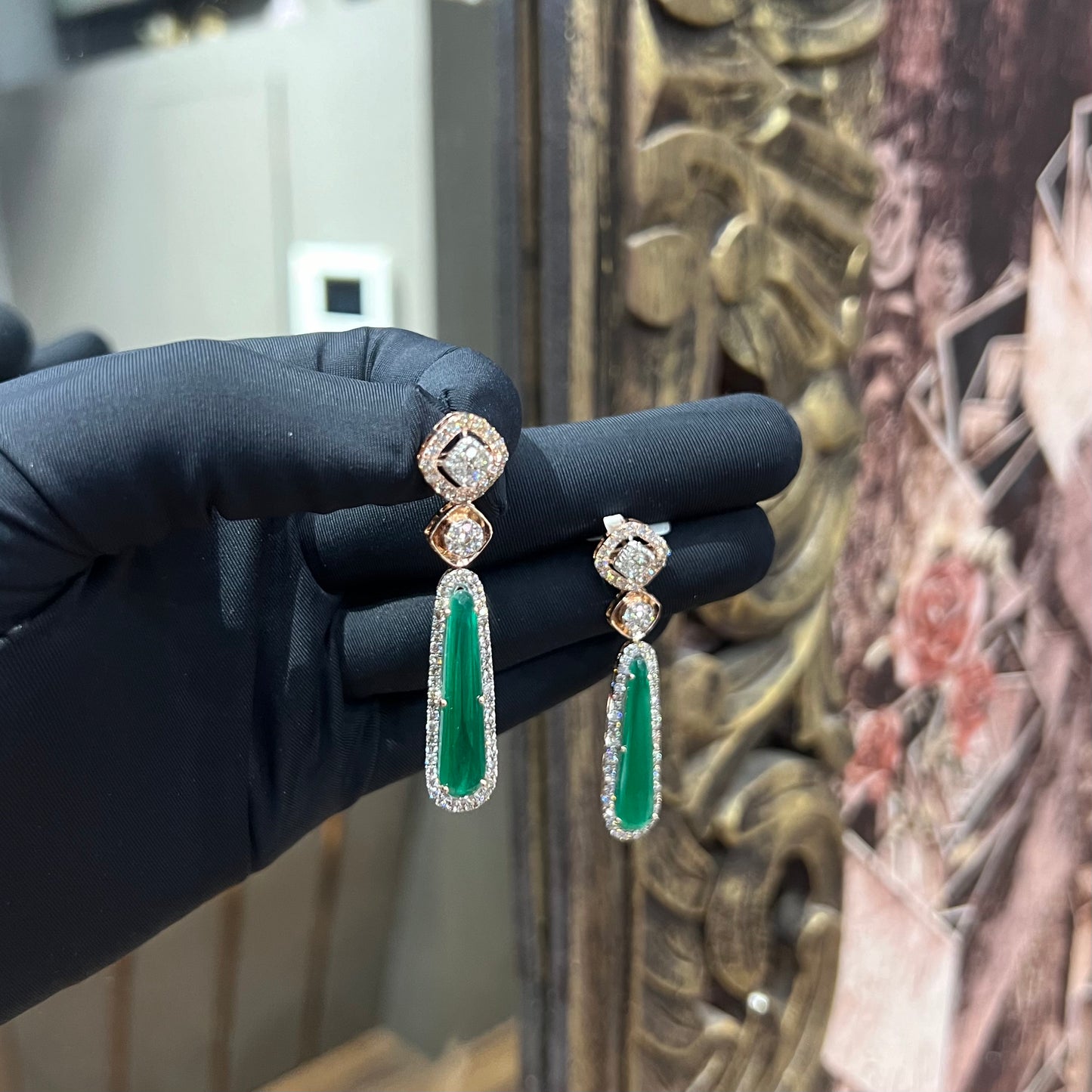 A pair of gold diamond earrings with green color stones set in a minimalist design.