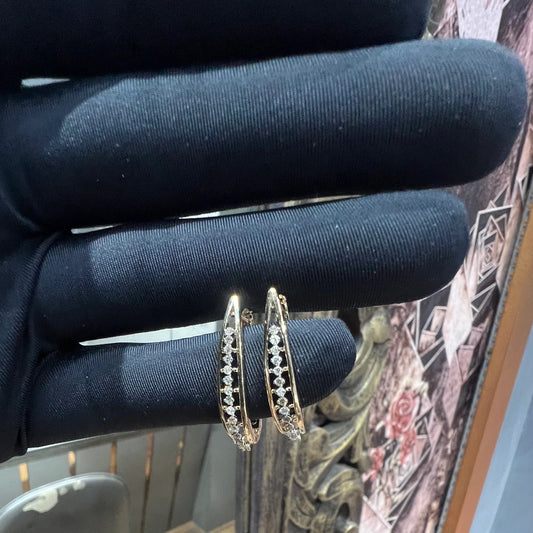 A pair of round-cut diamond hoop earrings held in a person's hand, with a gold band.