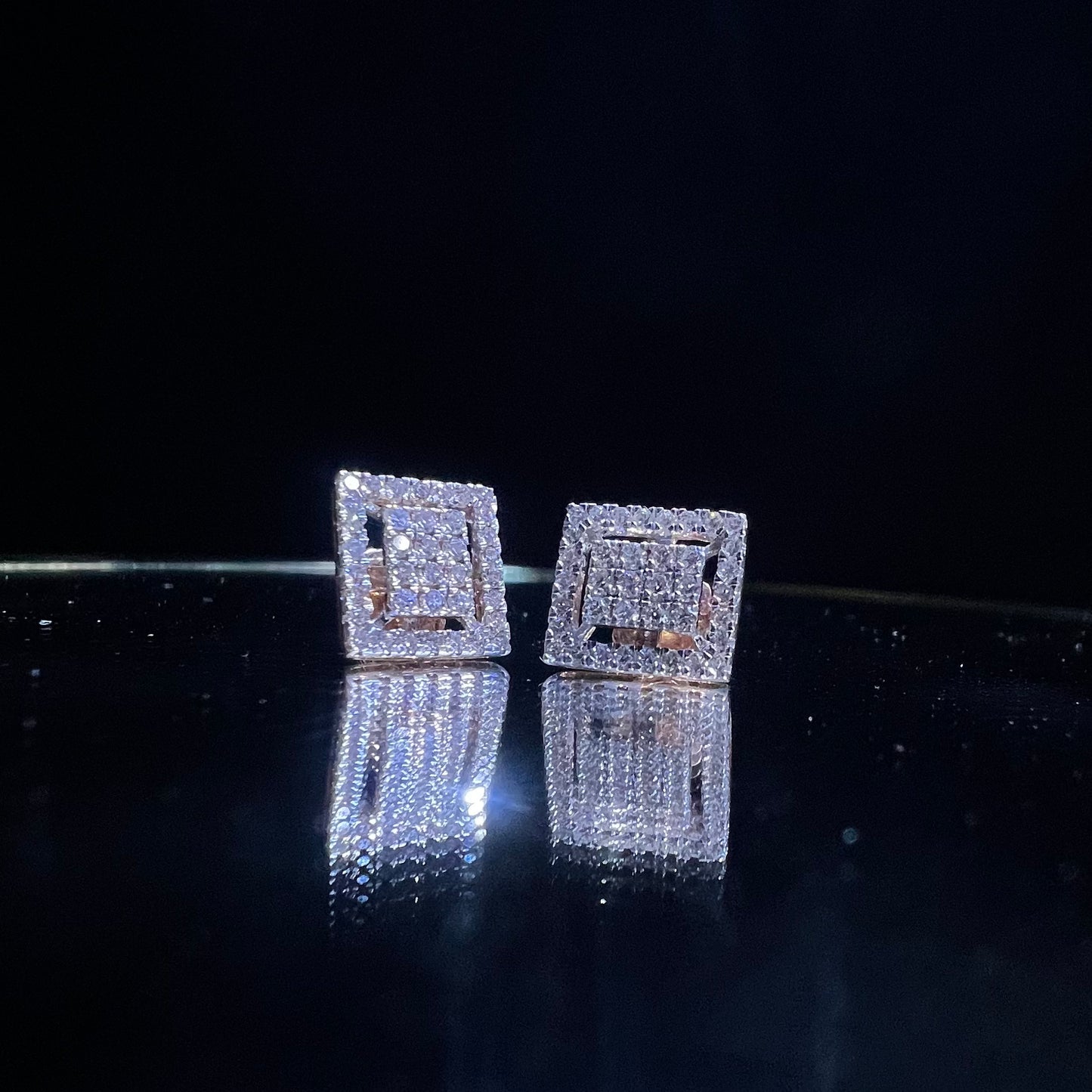 A pair of diamond stud earrings set in a gold casing, displayed against a reflective surface.