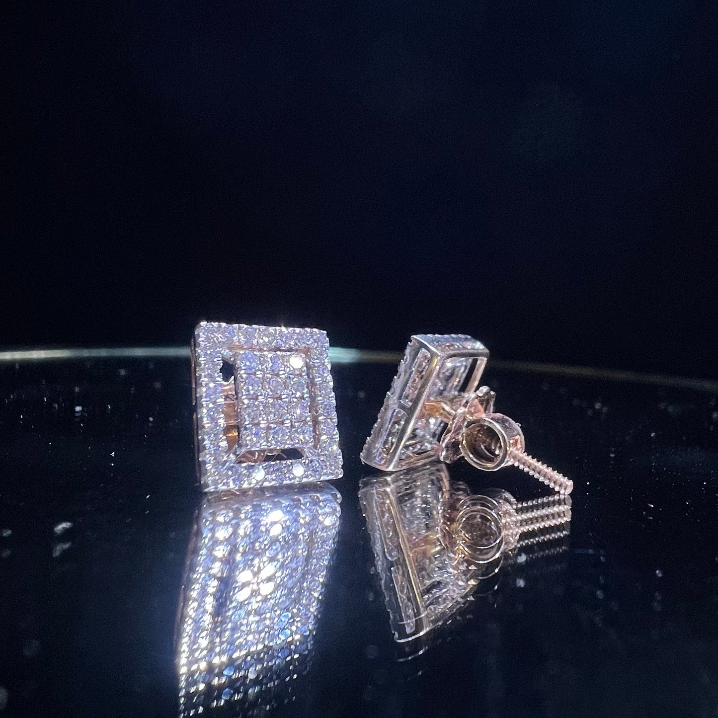A pair of diamond stud earrings set in a gold casing, displayed against a reflective surface.