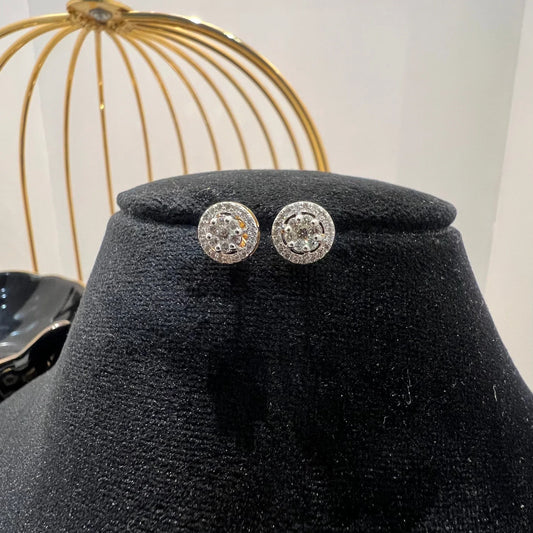 A pair of gold diamond earrings with a round cut design, placed on a mannequin stand with a decorative background.