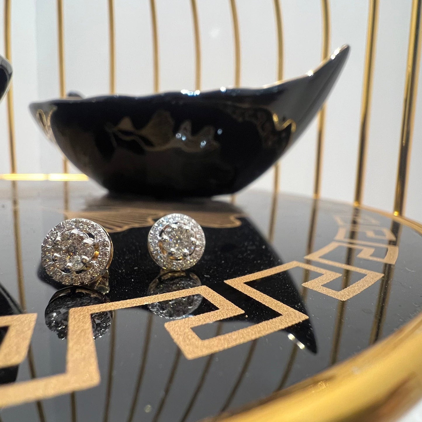A pair of gold diamond earrings with a round cut design, placed on a reflective surface with a decorative background.