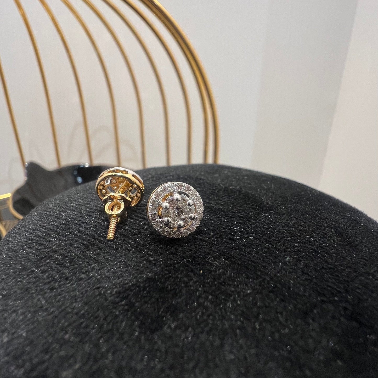 A pair of gold diamond earrings with a round cut design, placed on a mannequin stand with a decorative background.
