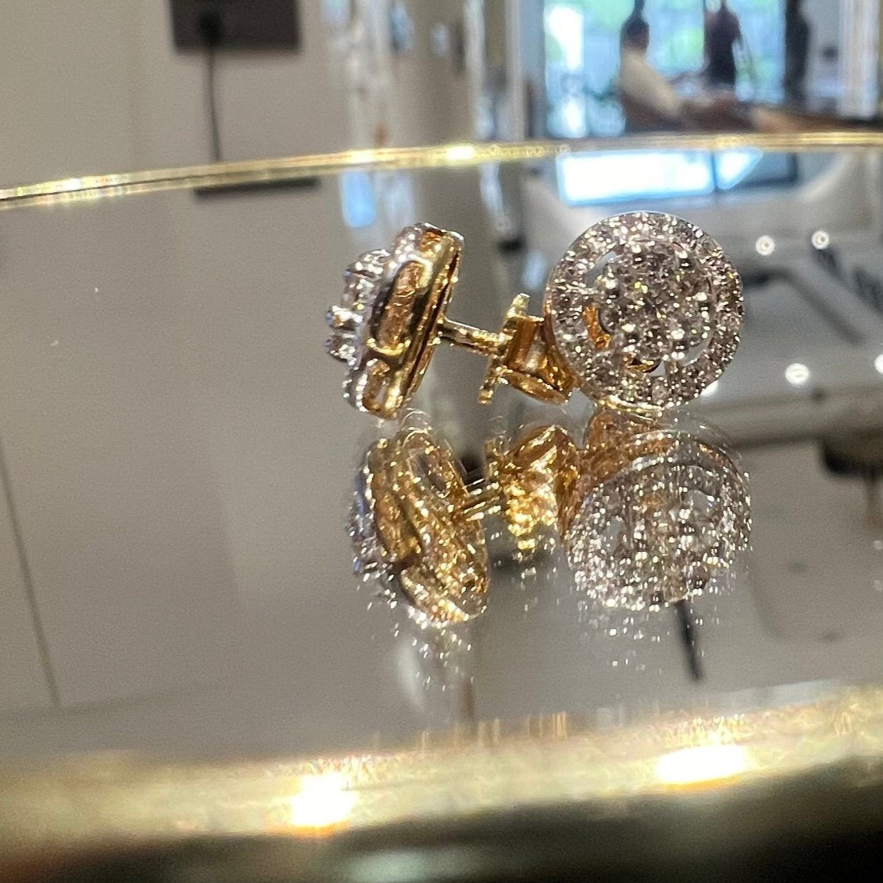 A pair of gold diamond earrings with a round cut design, placed on a reflective surface with a decorative background.