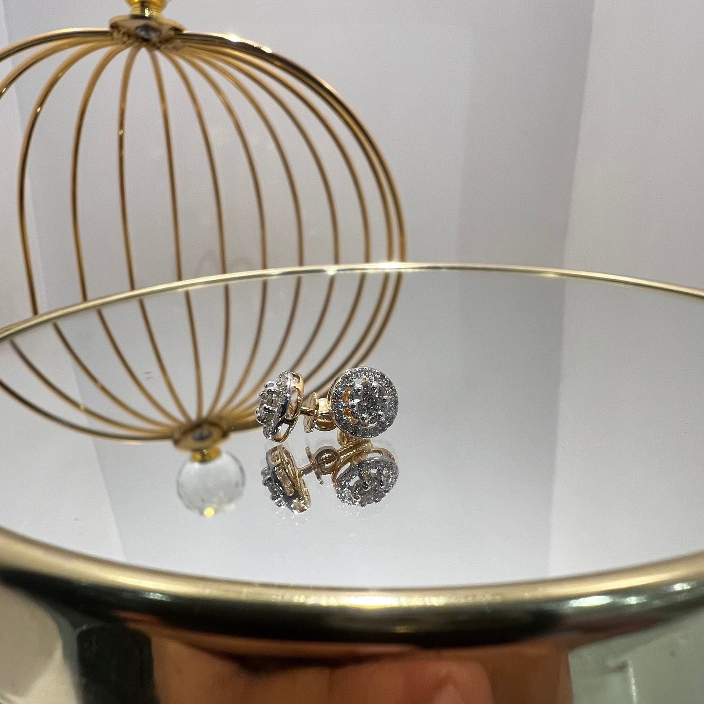 A pair of gold diamond earrings with a round cut design, placed on a reflective surface with a decorative background.