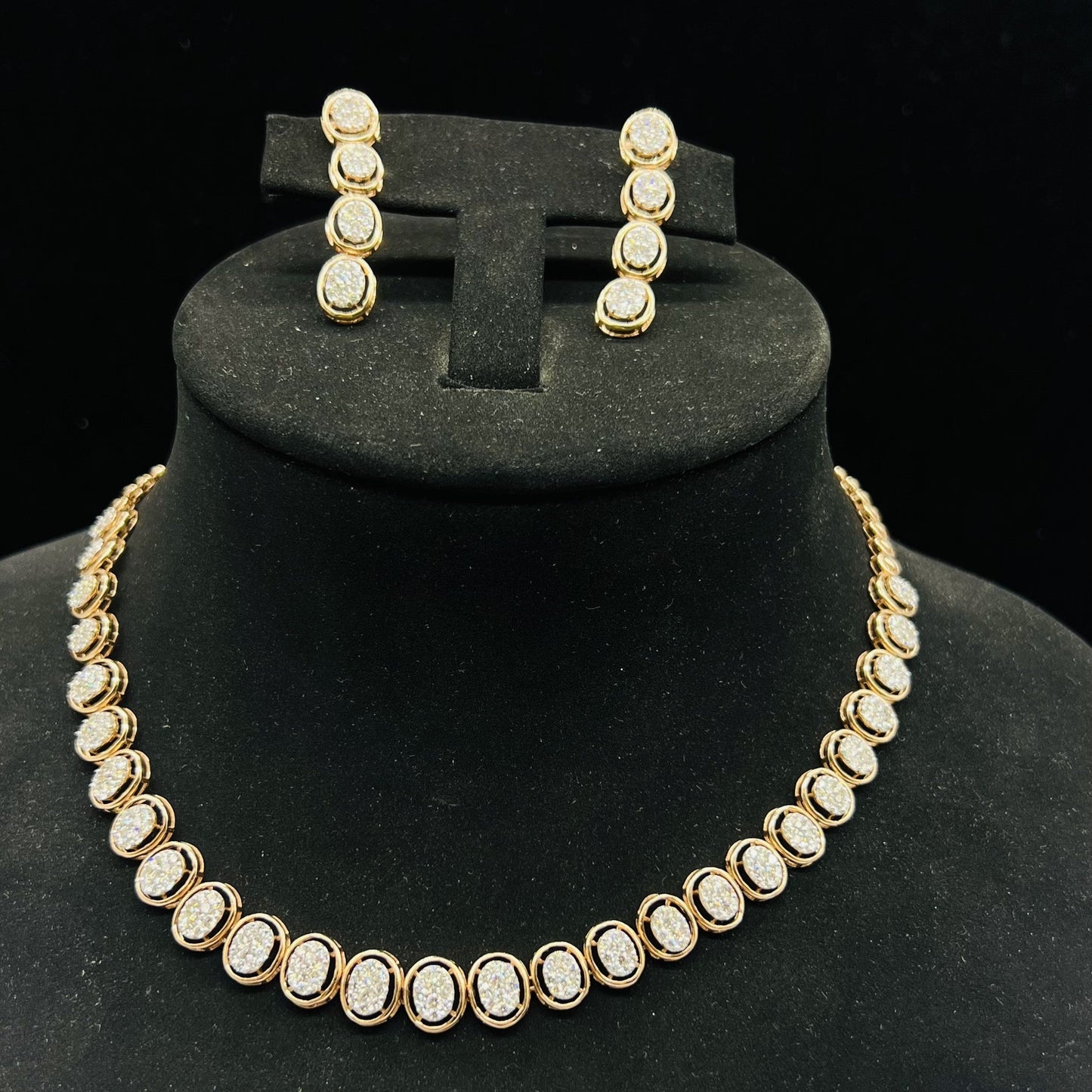 Gold diamond necklace set with matching earrings featuring oval-shaped, pavé-set diamonds in rose gold. The necklace has a linked design showcasing these sparkling stones evenly spaced. Earrings mirror the necklace stones in a vertical stack, creating an elegant accessory set on a black velvet background.