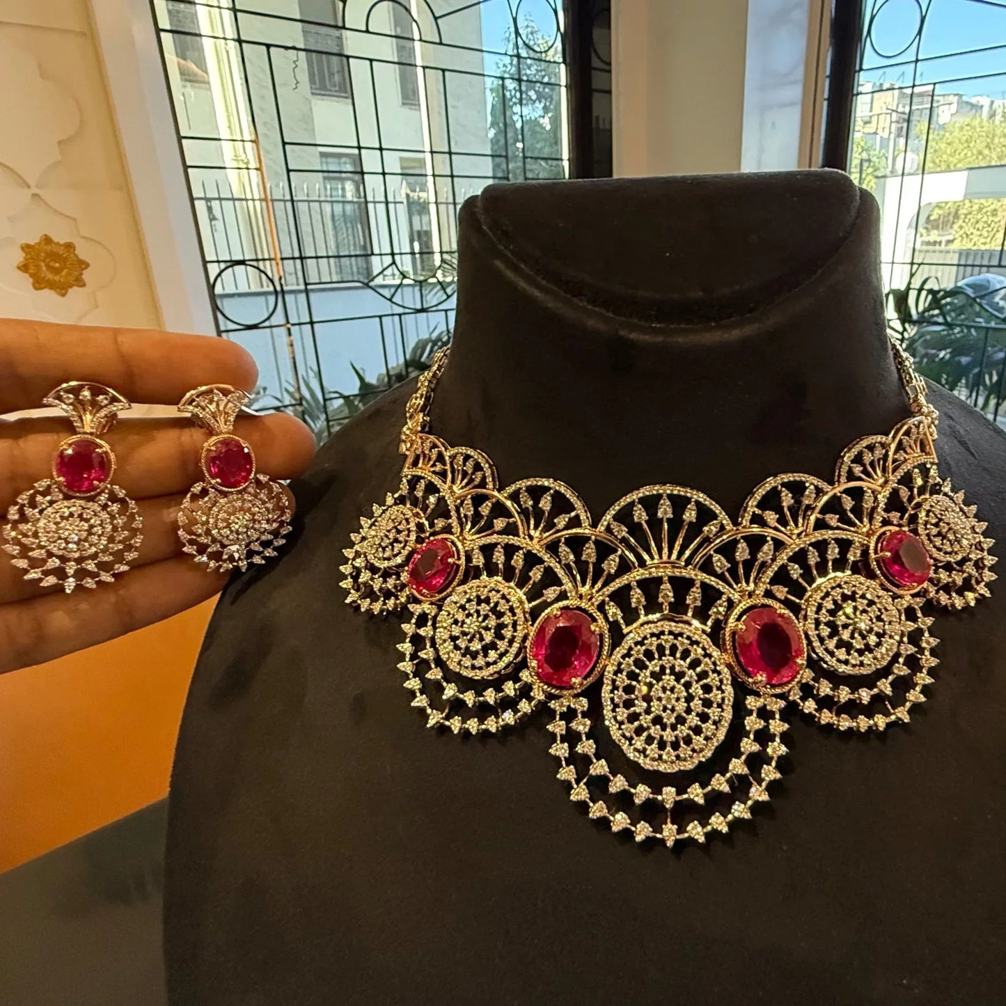 A gold diamond choker necklace with a floral pattern and red and white diamonds, displayed on a mannequin and accompanied by matching earrings on a hand.