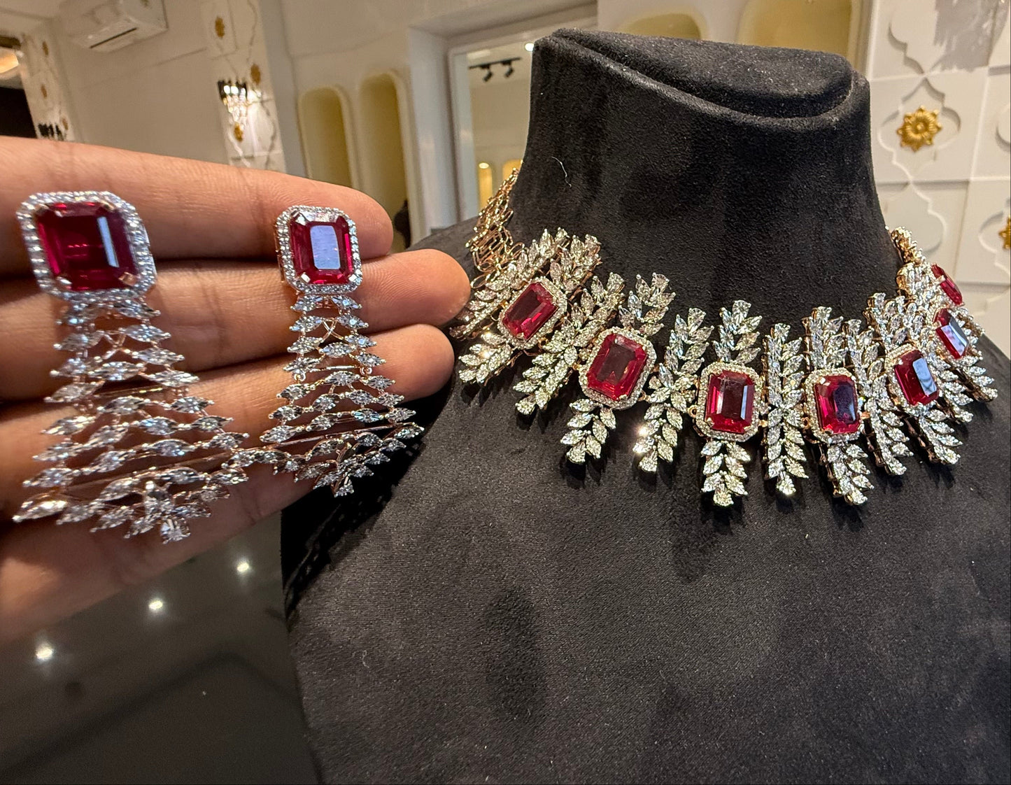 Diamond necklace set with emerald-cut color stones and leaf-shaped diamond clusters is displayed on a mannequin and matching color stone and diamond earrings are displayed on a hand.