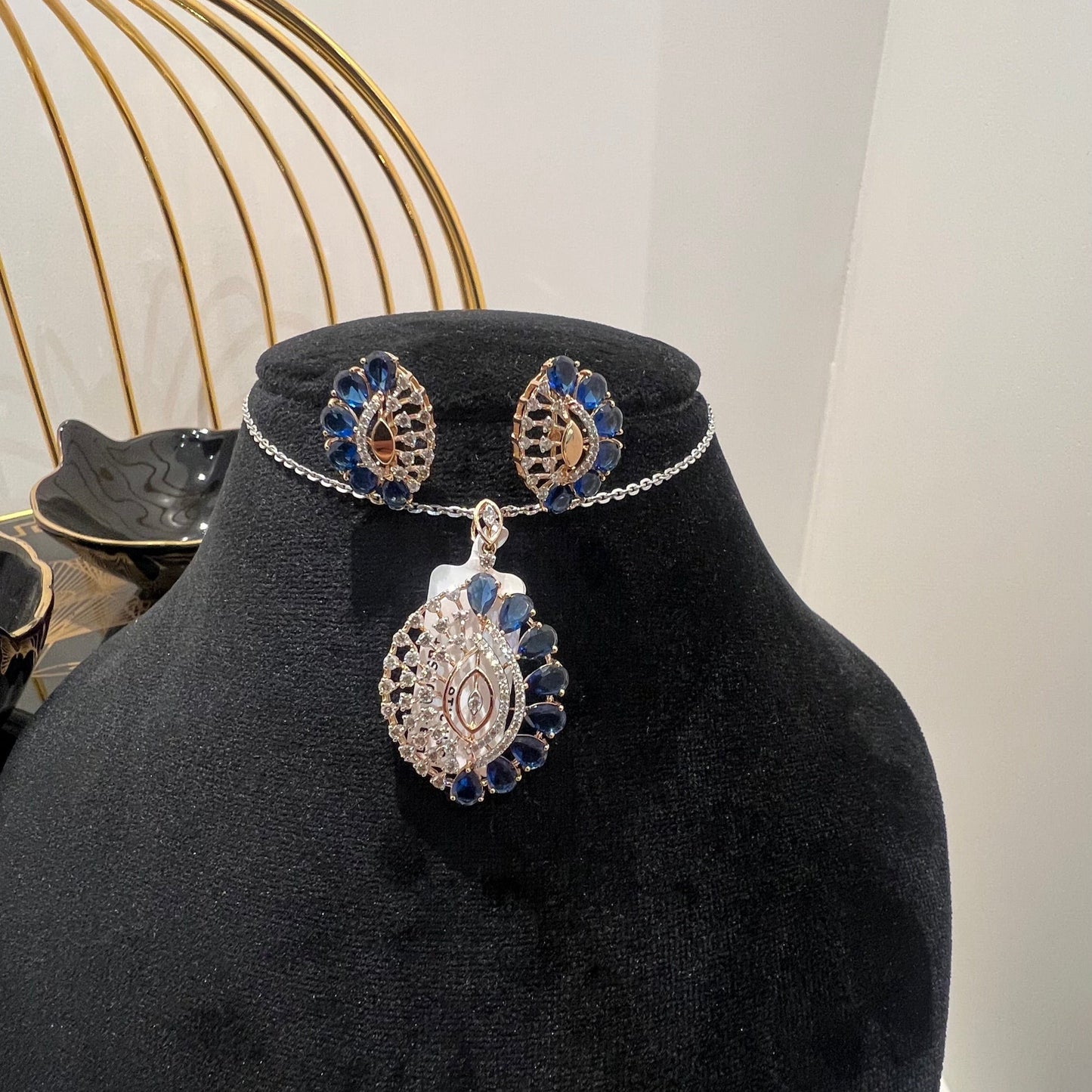 A gold diamond pendant with blue stone accompanied by matching earrings, displayed on a black mannequin bust.