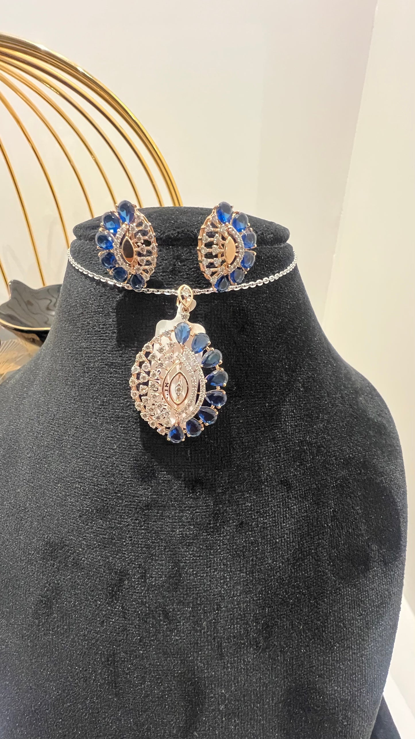 A gold diamond pendant with blue stone accompanied by matching earrings, displayed on a black mannequin bust.