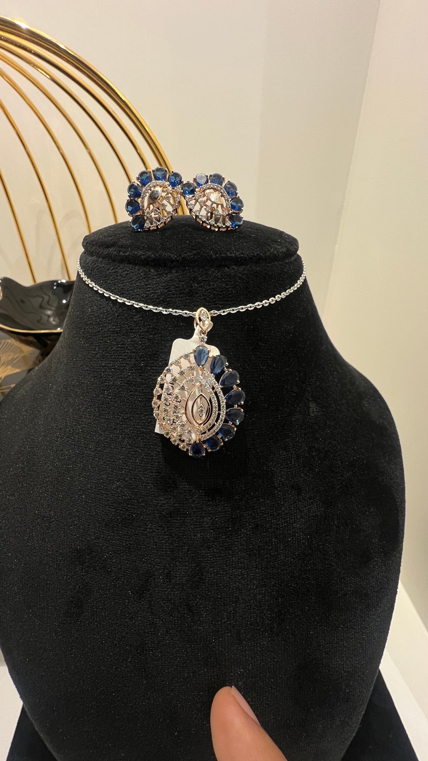 A gold diamond pendant with blue stone accompanied by matching earrings, displayed on a black mannequin bust.