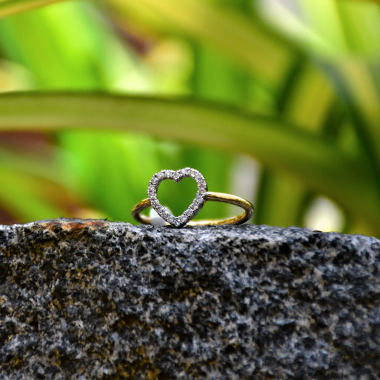 Delicate heart-shaped diamond ring set on a thin gold band, placed on a rocky surface against green and yellow variegated leaves in natural light.