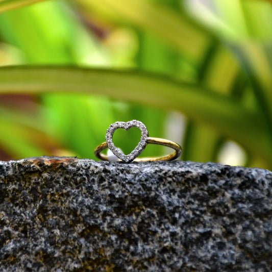 Delicate heart-shaped diamond ring set on a thin gold band, placed on a rocky surface against green and yellow variegated leaves in natural light.