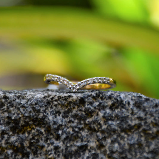 A gold ring featuring a V-shaped diamonds centerpiece, set on a rocky surface with a blurred green background.