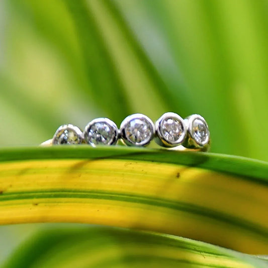 A yellow gold band ring with a series of set diamonds, featuring smaller diamonds, all set in a solid gold band displayed on a light green and yellow leaf.