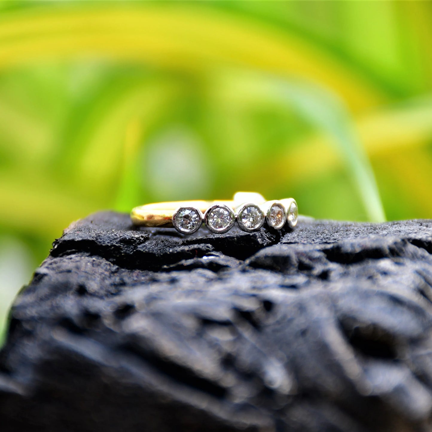 A yellow gold band ring with a series of set diamonds, featuring smaller diamonds, all set in a solid gold band, displayed against a light yellow and green background and on a black rough surface.