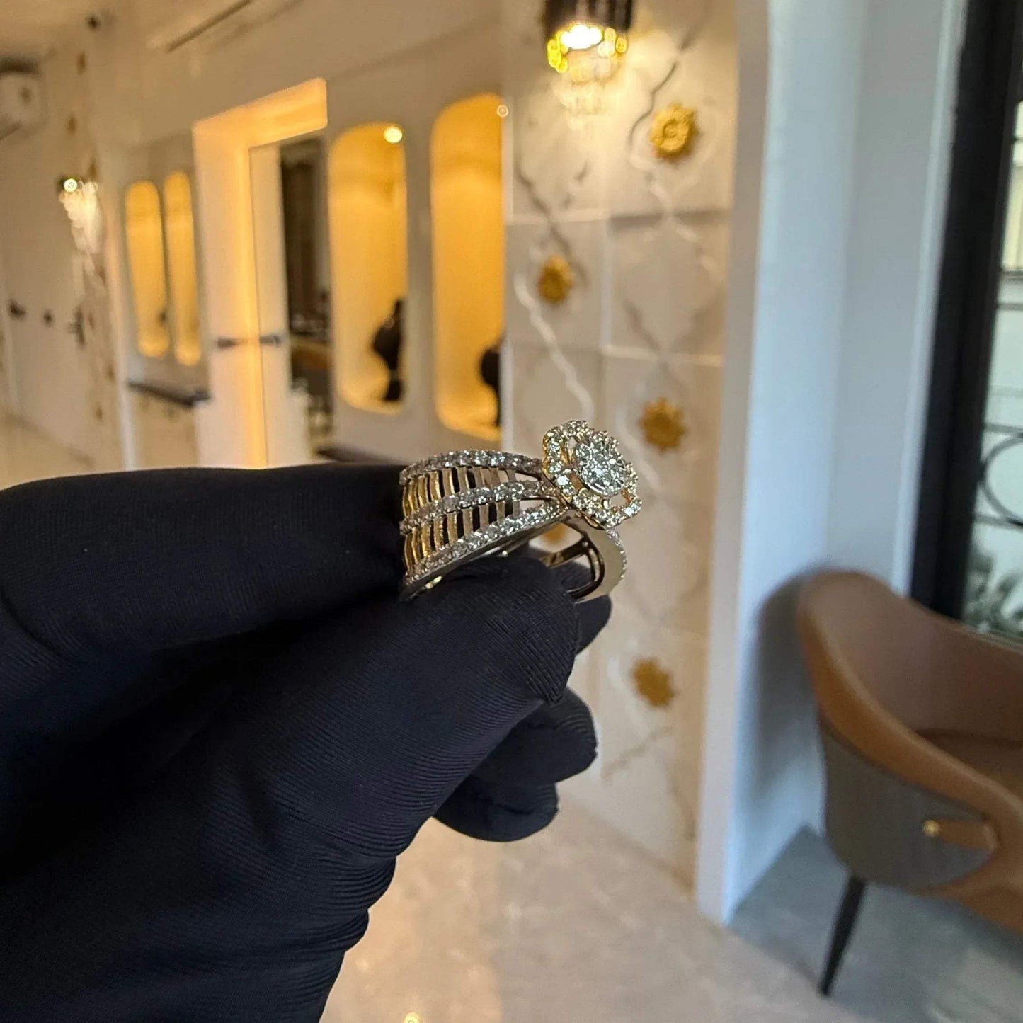 Elegant gold diamond ring featuring a round-cut central diamond surrounded by a halo of smaller diamonds, set on a split shank band adorned with rows of sparkling pavé diamonds, displayed on a black gloved hand against a neutral background.