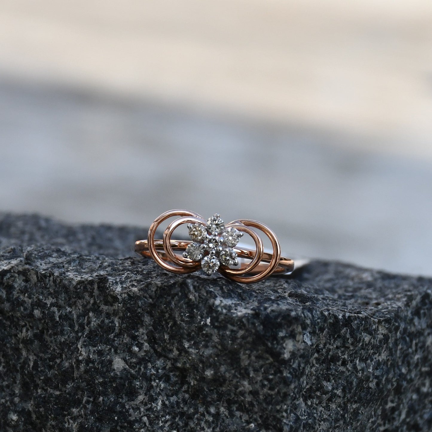 Delicate rose gold ring featuring a floral cluster of round diamonds at the center, showcased on a rough & textured surface.