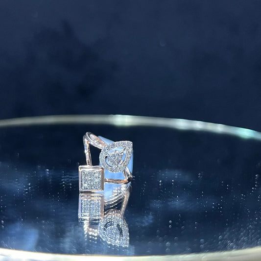 Elegant open-style diamond ring featuring a pear-shaped halo on one side and a square cluster on the other, displayed on a reflective metallic surface.