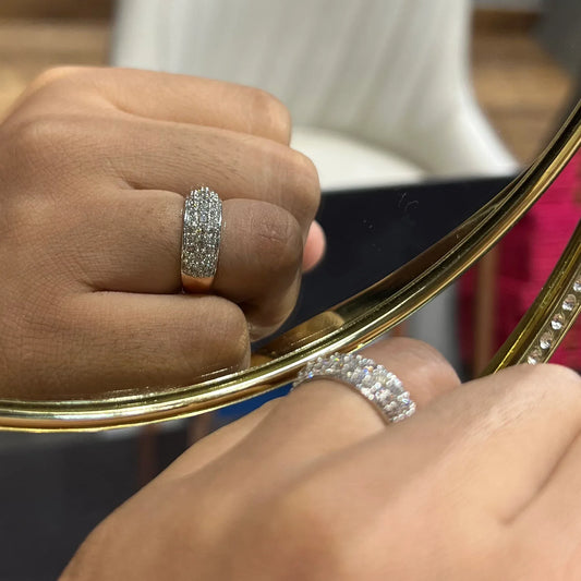Close-up of a hand wearing a wide diamond band ring, featuring multiple rows of sparkling round-cut diamonds set in gold, reflected elegantly in a gold-framed mirror.