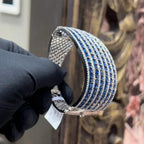 A close-up image of a hand holding a luxurious gold diamond bracelet with a sparkling blue stone, showing an openable design.