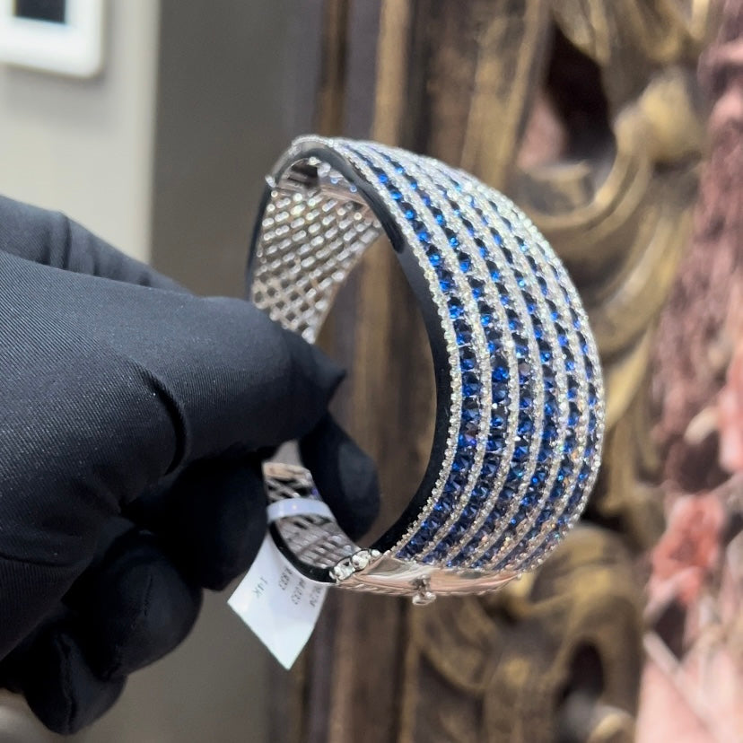 A close-up image of a hand holding a luxurious gold diamond bracelet with a sparkling blue stone, showing an openable design.