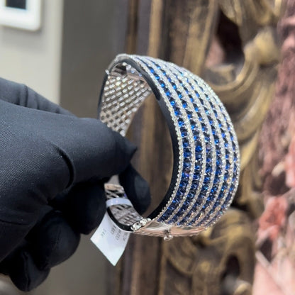 A close-up image of a hand holding a luxurious gold diamond bracelet with a sparkling blue stone, showing an openable design.