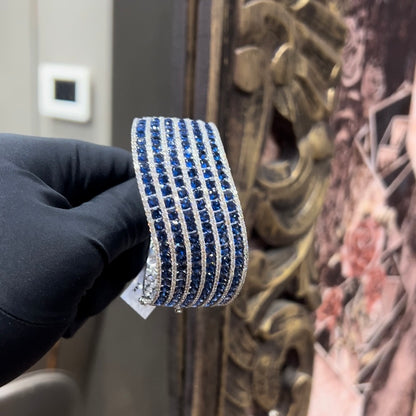 A close-up image of a hand holding a luxurious gold diamond bracelet with a sparkling blue stone, showing an openable design.