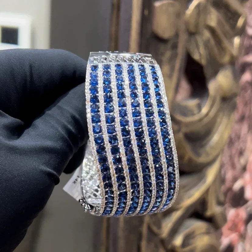 A close-up image of a hand holding a luxurious gold diamond bracelet with a sparkling blue stone, showing an openable design.