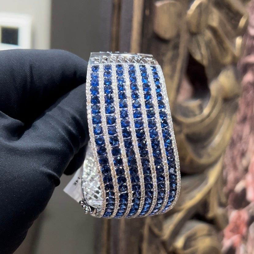 A close-up image of a hand holding a luxurious gold diamond bracelet with a sparkling blue stone, showing an openable design.