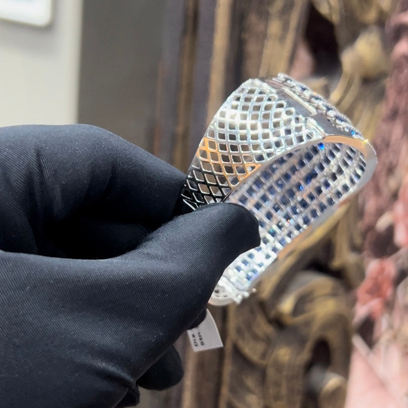 A close-up image of a hand holding a luxurious gold diamond bracelet with a sparkling blue stone, showing an openable design.