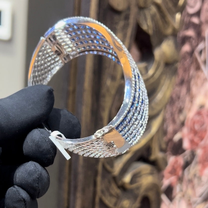 A close-up image of a hand holding a luxurious gold diamond bracelet with a sparkling blue stone, showing an openable design.