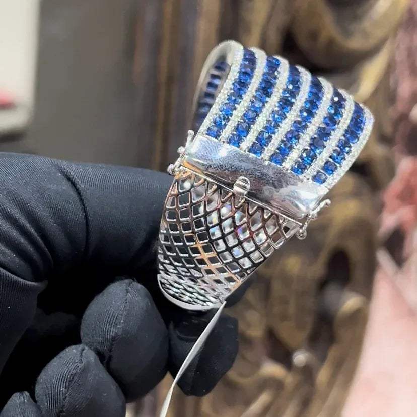 A close-up image of a hand holding a luxurious gold diamond bracelet with a sparkling blue stone, showing an openable design.