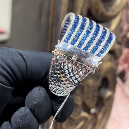 A close-up image of a hand holding a luxurious gold diamond bracelet with a sparkling blue stone, showing an openable design.
