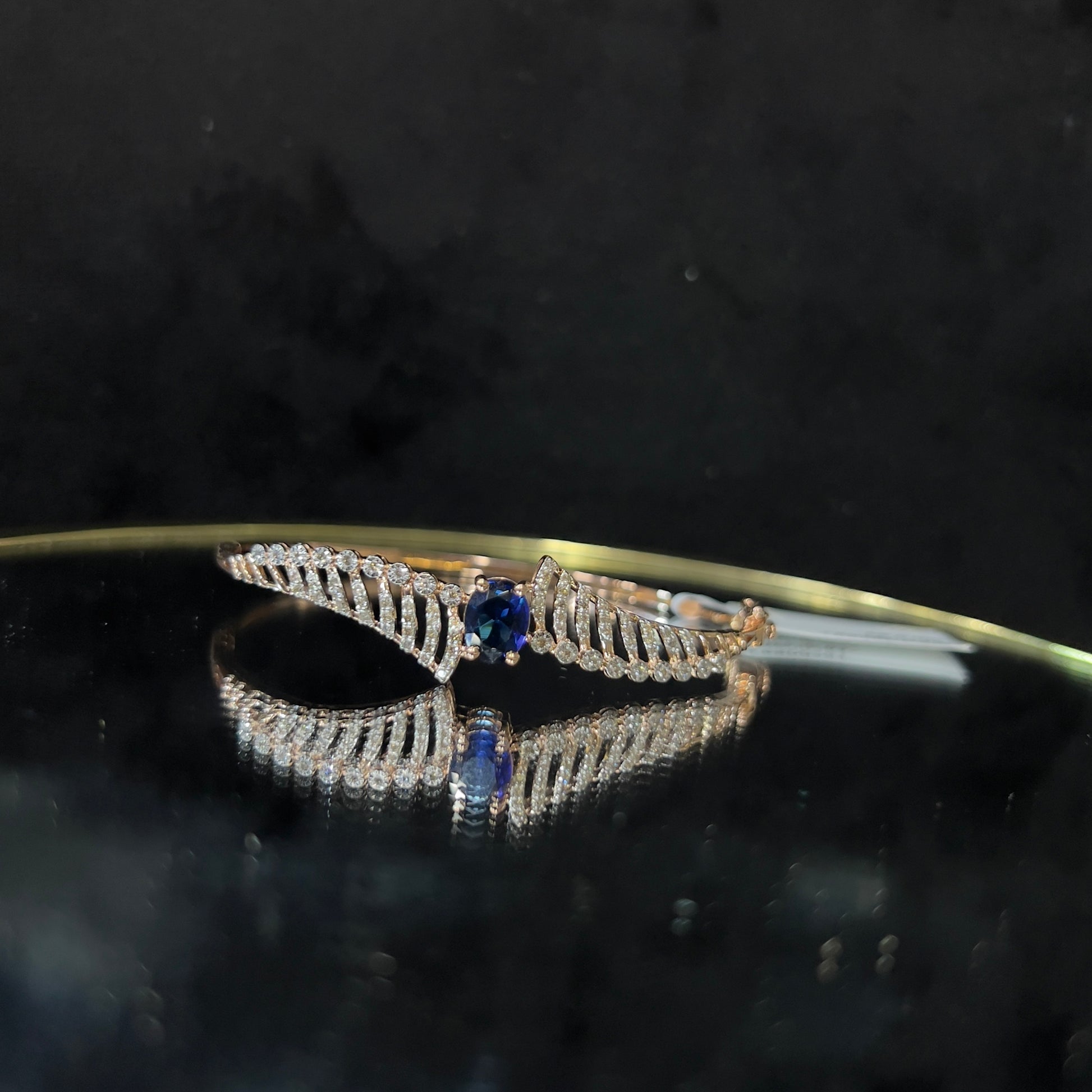 A luxurious gold diamond bracelet with a brilliant cut blue stone in an openable design, displayed on a reflective surface.