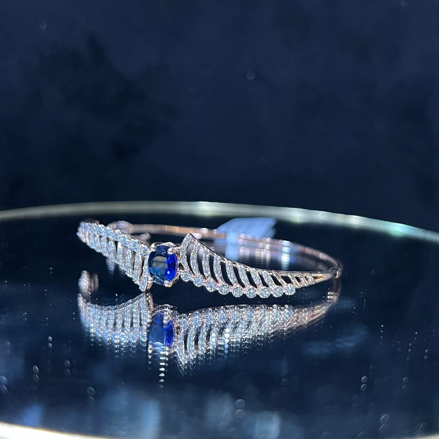 A luxurious gold diamond bracelet with a brilliant cut blue stone in an openable design, displayed on a reflective surface.